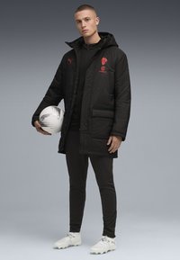 Sort puffer-jakke med rødt logo, båret over et sort basislag. Slanke sorte bukser og hvide fodboldstøvler, holder en sort og hvid fodbold.