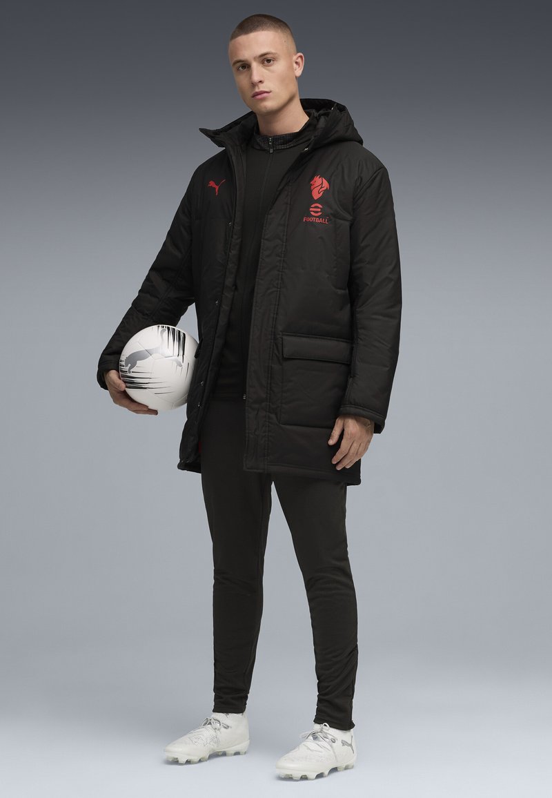 Sort puffer-jakke med rødt logo, båret over et sort basislag. Slanke sorte bukser og hvide fodboldstøvler, holder en sort og hvid fodbold.