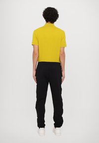 Achteraanzicht van een mannelijk model in een gele poloshirt, zwarte joggingbroek en witte sneakers. Strakke textuur, ontspannen pasvorm en eenvoudig ontwerp.