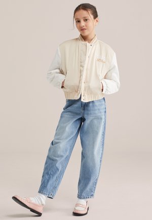 Deklica stoji z rokami v žepih, oblečena v bež bomber jakno, svetlo modre kavbojke, bele nogavice in roza loaferje na enobarvnem ozadju.