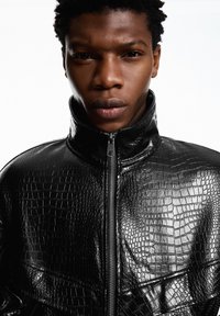 Zwarte jas van kunstleer met een glanzende krokodillenstructuur, voorzien van een hoge kraag en een ritssluiting aan de voorkant.