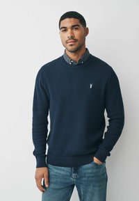 Granatowy sweter z dzianiny o żeberkowanej strukturze, okrągłym dekolcie i haftowanym logo na piersi, połączony z jasnoniebieskimi dżinsami.