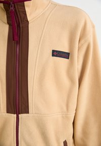 Beige fleece jacket featuring maroon zippers and side panels. A small black Columbia logo on the chest. Soft texture and stand-up collar.