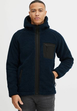 Navy blue fleece jacket with a hood, black zippered chest pocket, and hand pockets; textured surface, front zipper closure, and elastic cuffs.