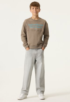 Sudadera gris con texto azul que dice "Inspired Illusions Surreal Sports", combinada con vaqueros gris claro y zapatillas blancas.