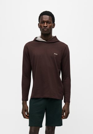 Uomo che indossa una felpa con cappuccio marrone scuro a maniche lunghe con il logo "BOSS" sul petto e pantaloncini verde scuro, in piedi davanti a uno sfondo chiaro e uniforme.