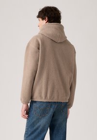 Tan fleece jacket with a hood, featuring a textured, plush surface and relaxed fit. Paired with blue denim jeans.