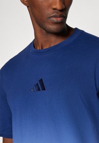 Modrá bavlnená tričká s klasickým výstrihom a krátkymi rukávmi, s tmavomodrým logom Adidas na hrudi. Hladký povrch.