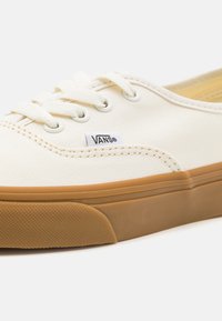 Baskets en toile blanche avec une semelle en caoutchouc beige, dotées d'œillets métalliques ronds et de lacets plats. Logo Vans brodé sur le côté.