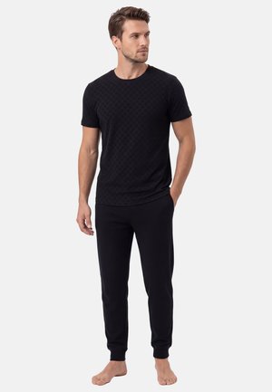 Homme debout pieds nus portant une chemise noire ajustée à manches courtes à motifs et un pantalon de jogging noir, regardant vers sa gauche.