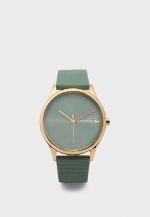 Goudkleurig horloge met een groene, gestructureerde siliconen band, een groen wijzerplaat, gouden wijzers en het Lacoste-logo aan de rechterkant van de wijzerplaat.
