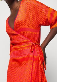 Ljust orange omslagsklänning med varierande mönster i rosa. Tillverkad av lätt tyg, med knyt i midjan och vida ärmar.