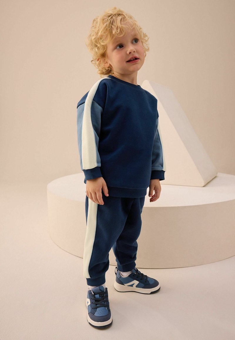 Sweatshirt en coton bleu foncé avec des rayures bleu clair et crème, associé à un jogging assorti et des sneakers bleus, se tenant sur une plateforme circulaire claire.