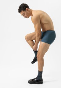 Homme portant un boxer à motifs bleu foncé, en train d'enfiler des chaussettes grises foncées et des chaussures noires, debout contre un fond blanc.