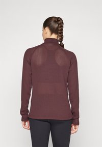 Bordó hosszúujjú felső textúrált mintával, magas gallérral és hálós részletekkel a hátsó részen; fekete leggings-szel párosítva.