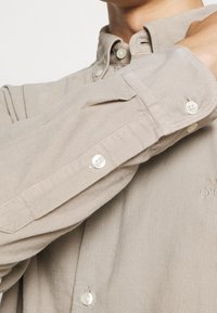 Camisa de pana beige con mangas largas, cuello abotonado y botones blancos. Presenta una textura acanalada y un logo bordado sutil.