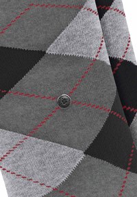 Des chaussettes argyle grises présentent un motif en losanges de nuances variées, avec des accents noirs et des coutures rouges. Un logo métallique circulaire est centré.