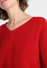 Pull en tricot rouge avec un décolleté en V, un tissu texturé et des manches larges. Agrémenté de boucles d'oreilles en or pendantes.
