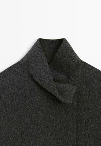 Tmavě šedý vlněný kabát s límcem s postaveným designem a prošívaným detailem zapínání na bílém pozadí.