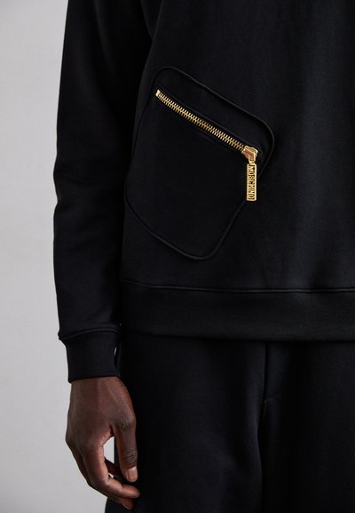 Čierna mikina s textúrovaným povrchom, ktorá má veľkú náprsné vrecko so zlatým zipsom. Sťahované rukávy a voľný strih.