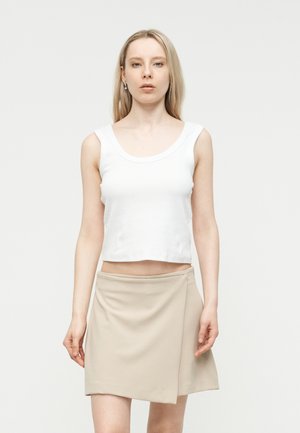 Junge Frau mit langen blonden Haaren, die ein weißes ärmelloses Oberteil und einen beigefarbenen Minirock trägt, vor einem hellen Hintergrund stehend.