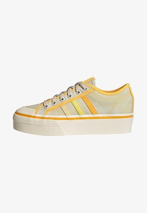 Zapatilla beige de caña baja con suela de plataforma, cordones grises y tres líneas laterales en amarillo y naranja.