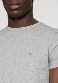 Primo piano di una persona che indossa una t-shirt grigia chiaro con un piccolo logo rettangolare rosso, bianco e blu sul petto.