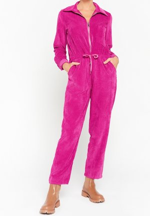 Kobieta w jasnoróżowym sztruksowym kombinezonie z suwakiem z przodu, ściągaczem w talii, bocznymi kieszeniami i beżowymi botkami za kostkę.