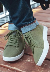Persona con zapatillas de punto verde oliva y suelas blancas, vestida con jeans oscuros y ajustados, de pie sobre tablones de madera.