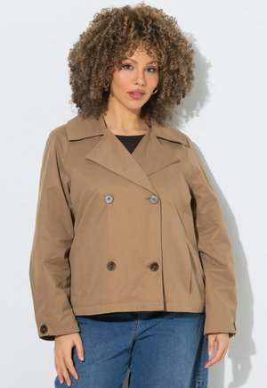 Kvinde med krøllet hår iført en beige dobbeltradet jakke og blå jeans, stående mod en ensfarvet hvid baggrund.