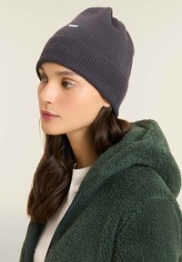 Berretto lavorato a maglia grigio scuro con una banda a costine e un piccolo logo bianco. Indossato con una giacca in pile verde, che mostra una texture morbida e uno stile informale.