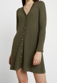 Robe en olive verte côtelée avec manches longues, col en V et boutons marron sur le devant, dotée d'un ourlet évasé et d'une texture douce.