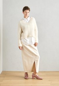 Un pull surdimensionné en crème en peluche porté par-dessus une chemise blanche à boutons, associé à une jupe fendue beige et des chaussures à talons hauts roses avec une touche dorée.