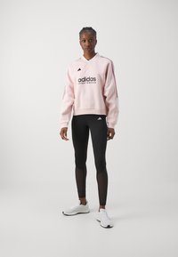 Roze sweatshirt met zwart logo en drie verticale strepen, gecombineerd met zwarte legging met mesh accenten en witte sportschoenen.