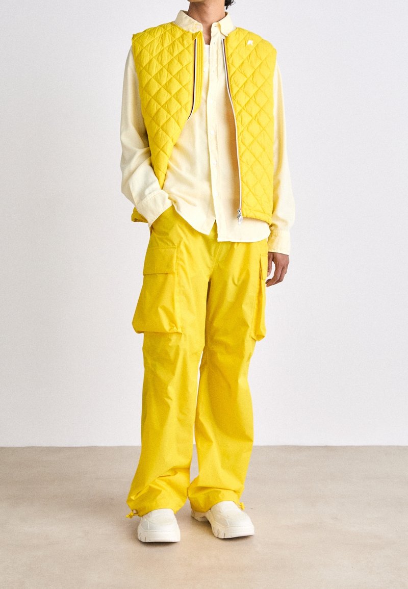 Persona che indossa un gilet trapuntato giallo, una camicia button-up giallo chiaro, pantaloni cargo giallo vivace e sneakers bianche, in piedi su un pavimento beige.