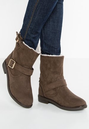 Bottes chelsea en daim marron avec boucle et sangle latérales, doublure en polaire blanche, portées avec un jean skinny bleu foncé.