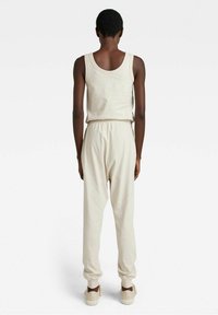 Person som står med ryggen mot kameran, iklädd en krämfärgad linne, matchande joggingbyxor och beige sneakers på en vit bakgrund.