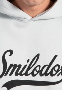 Felpa leggera grigia realizzata in tessuto morbido. Presenta un grande testo nero "Smilodox" sulla parte anteriore con una texture liscia e senza abbellimenti.