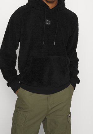 Mann trägt schwarzen, flauschigen Hoodie mit Fronttasche und olivgrüne Cargo-Hose, Hände in den Taschen, steht vor weißem Hintergrund.