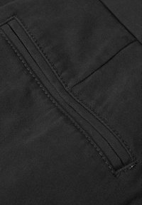 Gros plan sur un tissu noir avec une poche passepoil verticale et des coutures visibles sur le textile environnant.