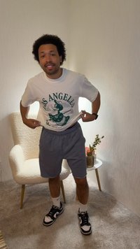 Νέος άντρας στέκεται σε ένα δωμάτιο σηκώνοντας ένα λευκό T-shirt με γραφικά του Λος Άντζελες, φοράει γκρι σορτς και ασπρόμαυρα αθλητικά παπούτσια.