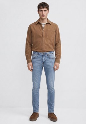 Junger Mann steht, trägt ein braunes Hemd mit Knöpfen über einem gestreiften T-Shirt, hellblaue Jeans und braune Freizeitschuhe vor neutralem Hintergrund.