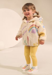 Veste en polaire couleur crème avec motifs arc-en-ciel multicolores et points, associée à des leggings jaunes ornés d'appliqués étoiles et des baskets colorées.
