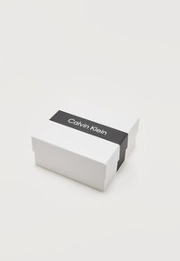 Boîte rectangulaire blanche avec une bande noire autour portant le logo Calvin Klein en texte blanc, sur un fond clair et neutre.