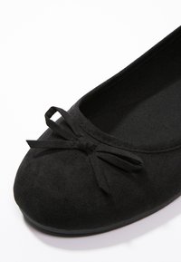 Ballerine en suede noir avec un bout arrondi, ornée d'un petit nœud sur le devant et d'une doublure intérieure texturée. Surface douce et lisse.