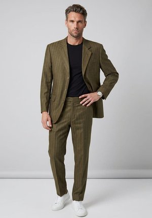 Hombre con traje verde oliva de rayas finas, con camisa negra y zapatillas blancas, de pie frente a un fondo gris liso.