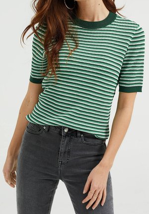 Woman wearing green and white striped short-sleeve knit top and dark gray high-waisted jeans, standing with one hand relaxed by side.