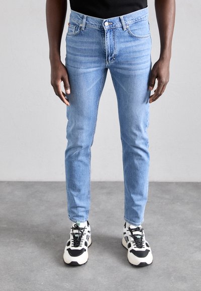 Világoskék, slim fit farmernadrág, öt zsebes dizájn és diszkrét kopásokkal. Fekete és fehér sportcipővel párosítva.