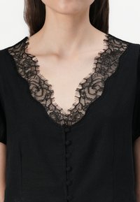 Indumento nero a maniche corte, caratterizzato da un profondo scollo a V ornato con delicata pizzo nero e piccoli bottoni in tessuto lungo la parte anteriore.