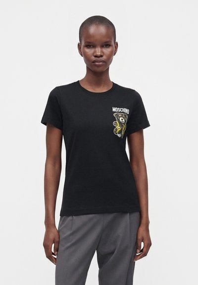Άτομο που φοράει μαύρο T-shirt Moschino με γραφικό αρκουδάκι και γκρι παντελόνι, στέκεται μπροστά σε απλό φωτεινό φόντο.